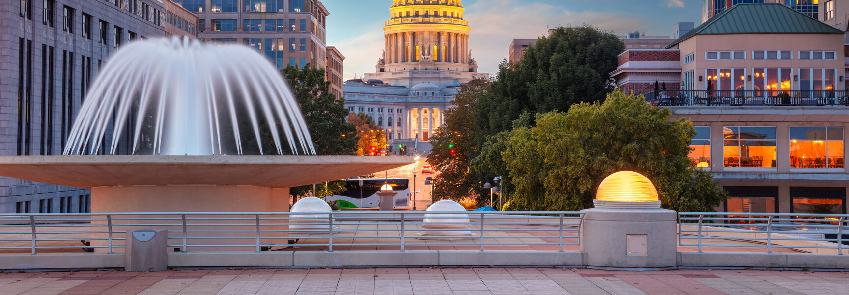 View of the illuminated Wisconsin State Capitol dome at sunset from a plaza with a large white fountain in Madison.