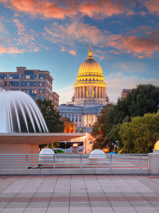 View of the illuminated Wisconsin State Capitol dome at sunset from a plaza with a large white fountain in Madison.