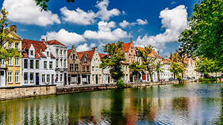 Historic gabled buildings line a calm canal in Bruges, Belgium, reflecting in the water under a bright sky with fluffy white clouds.