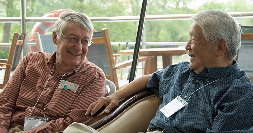 Two elderly male passengers wearing name tags smile and converse while sitting on the deck of a boat.