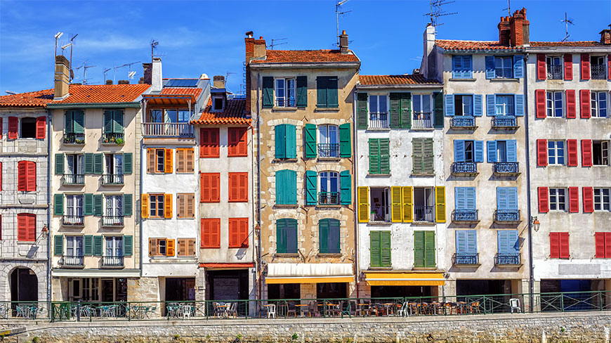 Colorful traditional Basque-style houses with vibrant shutters lining the waterfront in Bayonne, France, under a clear blue sky.