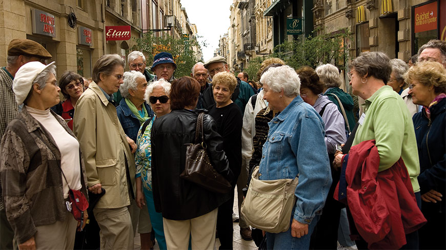 A group of senior tourists gathers on a busy pedestrian street lined with shops and historic architecture in Bordeaux, France.