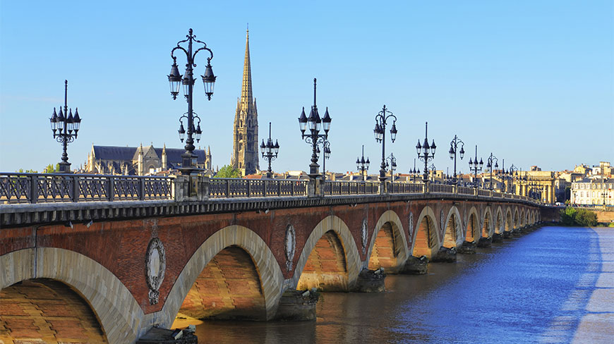 The multi-arched stone Pont de Pierre bridge in Bordeaux, France, features ornate street lamps and a cathedral spire in the background.