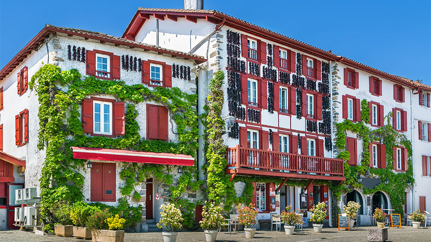 Traditional white buildings in Espelette, France, featuring red shutters, hanging dried peppers, and lush green vines under a clear blue sky.
