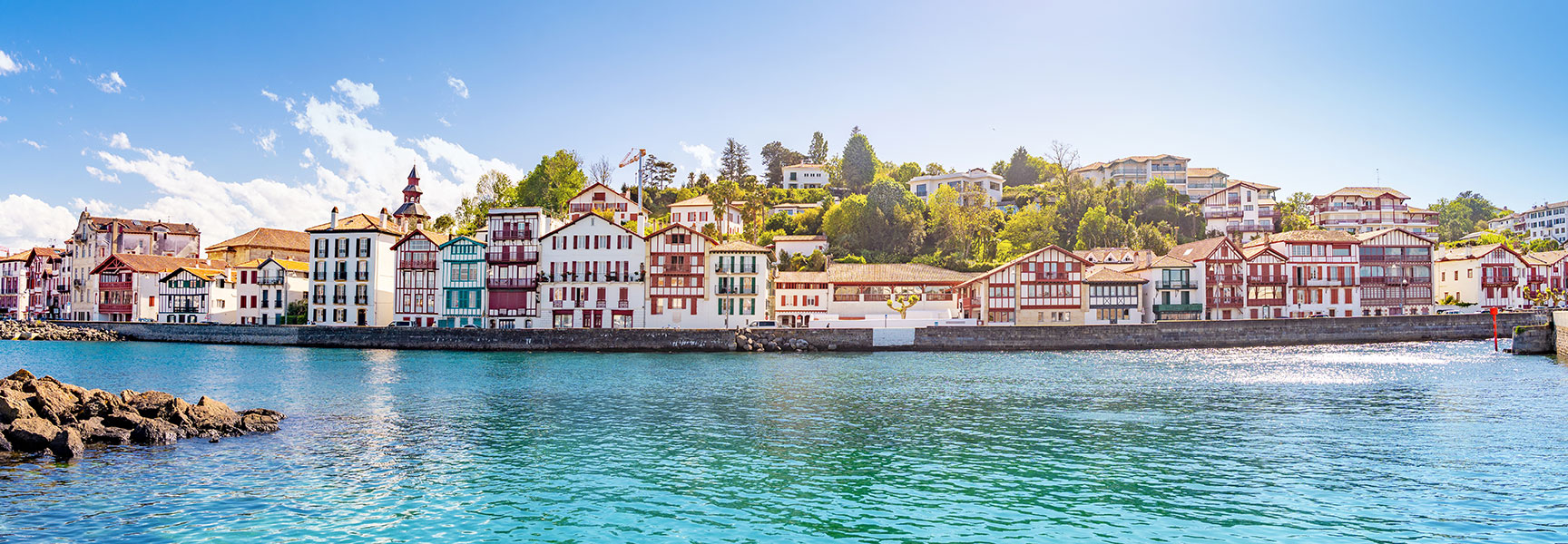 Panoramic view of colorful traditional Basque houses lining the harbor of Saint-Jean-de-Luz under a bright blue sky.