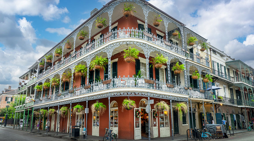 Ornate red building in the New Orleans French Quarter features multi-level wrought iron balconies adorned with lush hanging plants under a bright, cloudy sky.