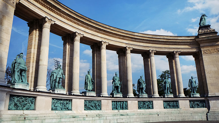 The curved colonnade of Heroes' Square in Budapest features bronze statues of historical figures between stone pillars under a clear blue sky.