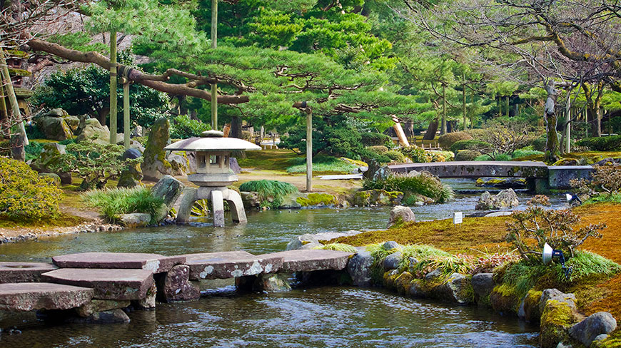 A stone lantern sits on the bank of a stream in a tranquil Japanese garden with stone bridges and pine trees.