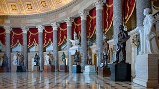 The ornate National Statuary Hall, a large semicircular room with marble columns, red drapery, a polished floor, and numerous statues of prominent figures.
