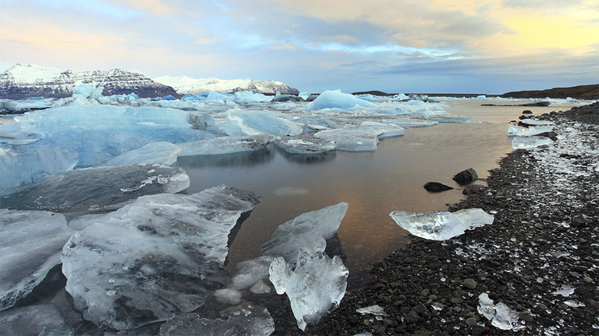 Large blue icebergs drift in the Jokulsarlon Glacier Lagoon in Iceland, surrounded by a dark pebble shore and distant snow-capped mountains at sunset.