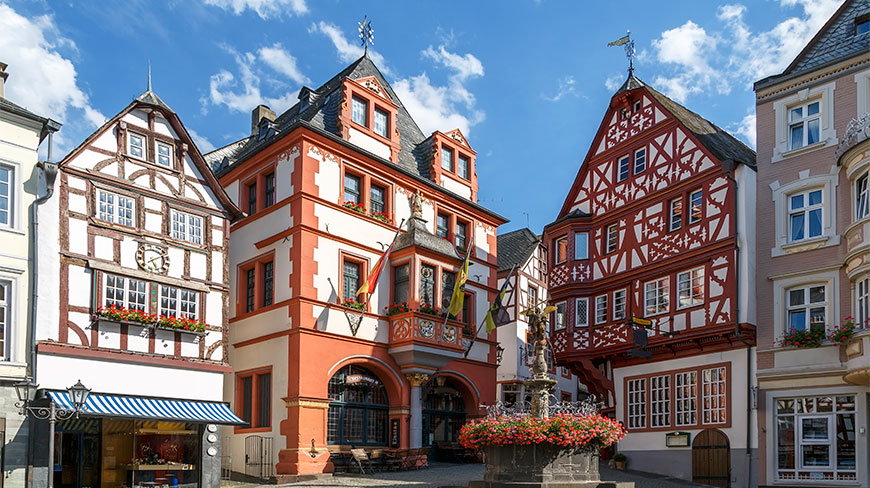 Colorful half-timbered buildings and a stone fountain decorated with flowers in the historic market square of Bernkastel-Kues, Germany.