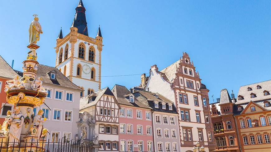 The historic Main Market in Trier, Germany, showcasing colorful architecture, the St. Gangolf church tower, and an ornate stone fountain against a bright blue sky.