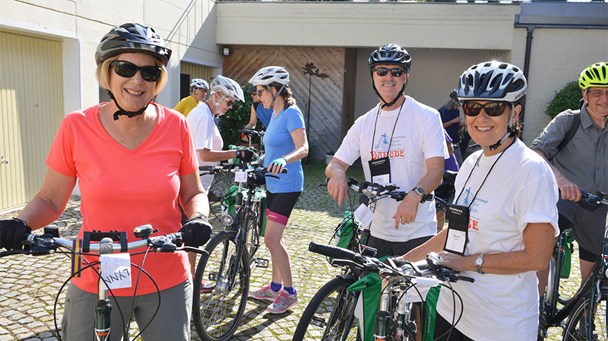 A smiling group of people wearing helmets and athletic attire stand with their bicycles outdoors, prepared for a DE biking group tour.
