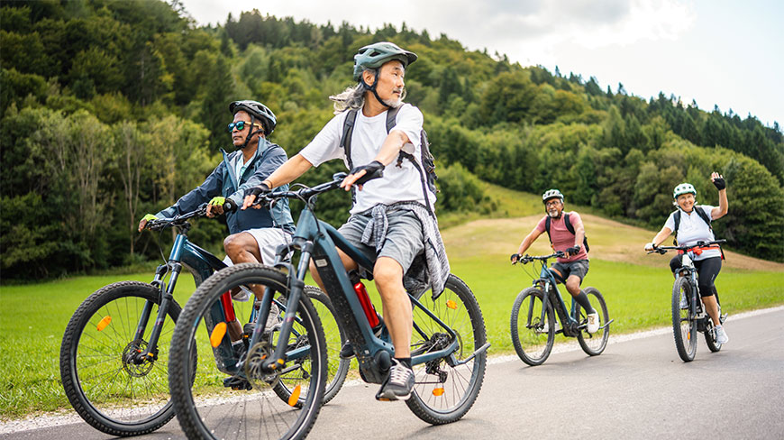 A group of diverse older adults biking on a paved road through a scenic green valley with forested hills in the background.