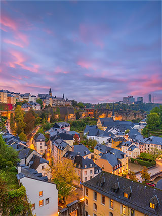A panoramic view of the Luxembourg City old town at twilight with glowing streetlights and a colorful sunset sky.