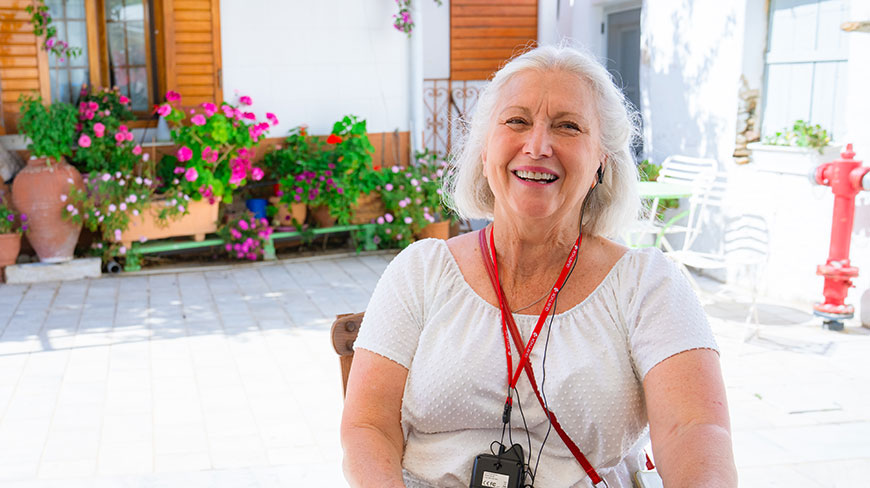 A smiling woman with white hair wears an audio guide lanyard during a walking tour in Lefkes, Paros, surrounded by colorful flowers.