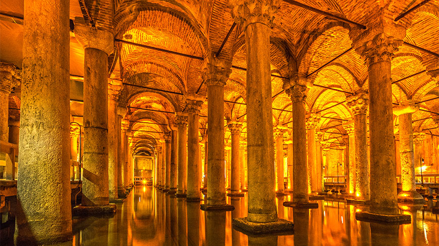 Warmly lit rows of ancient stone columns and arched brick ceilings reflecting in the still water of the Basilica Cistern in Istanbul.