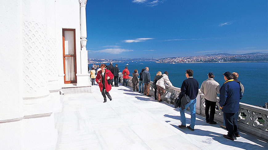 People on a white marble terrace overlooking the Istanbul sea and distant city buildings under a bright blue sky.