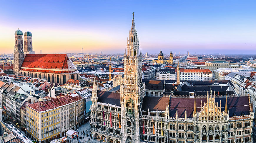 A panoramic view of the Munich city center featuring the historic New Town Hall and the iconic twin towers of the Frauenkirche at sunset.