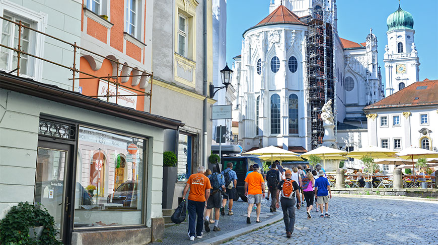 Tourists walk on a cobblestone street toward St. Stephen's Cathedral in Passau, Germany, passing colorful buildings and outdoor café umbrellas.