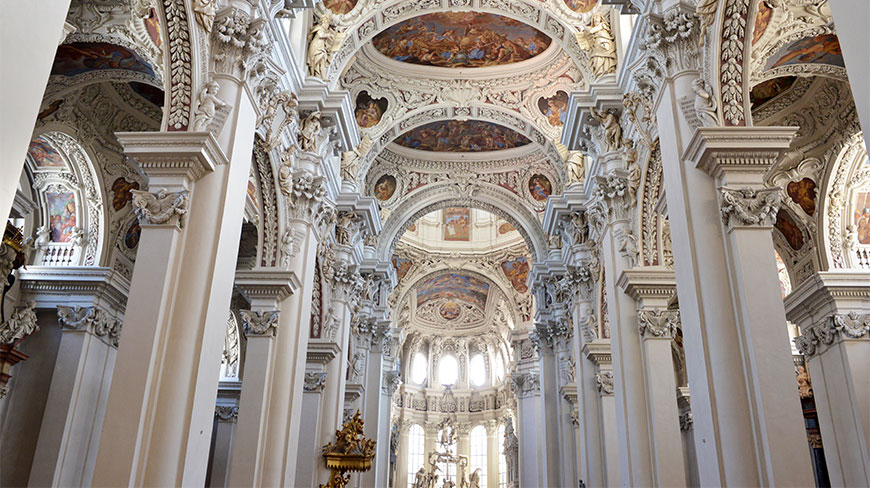 The ornate baroque interior of St. Stephen's Cathedral in Passau features white columns, intricate sculptures, and colorful ceiling frescoes.