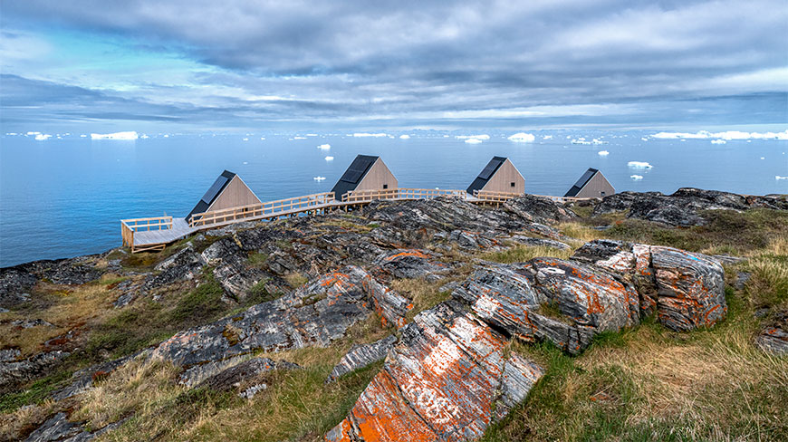Small modern wooden cabins sit on a rocky shoreline overlooking the iceberg-filled waters of the Illulissat Icefjord in Greenland.