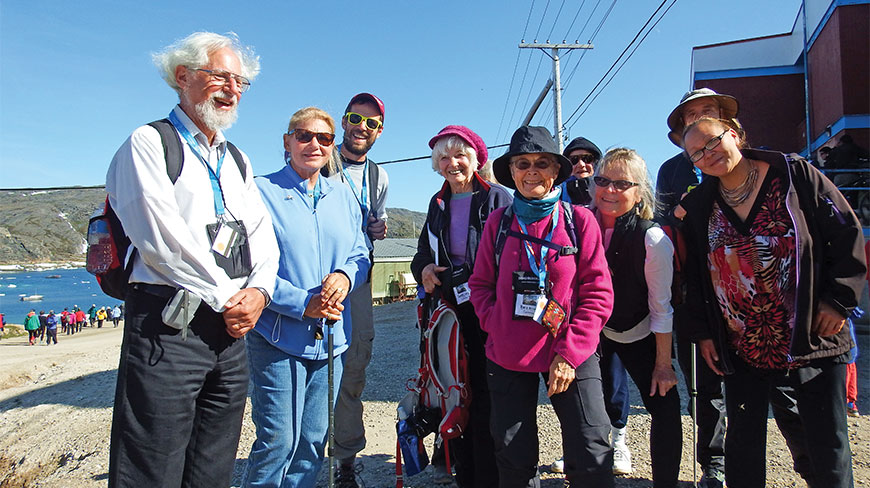 A group of smiling travelers stands outdoors in hiking gear during a Heart of Arctic expedition with water and ice visible in the background.