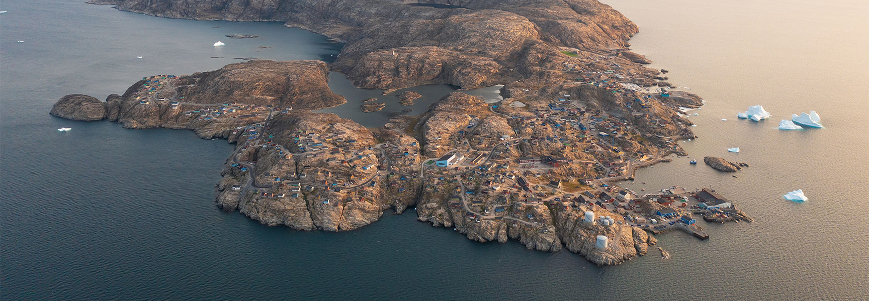 An aerial view of Uummannaq town in Greenland, featuring a large, heart-shaped mountain overlooking the rocky coastal settlement surrounded by icebergs.