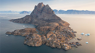 An aerial view of Uummannaq town in Greenland, featuring a large, heart-shaped mountain overlooking the rocky coastal settlement surrounded by icebergs.