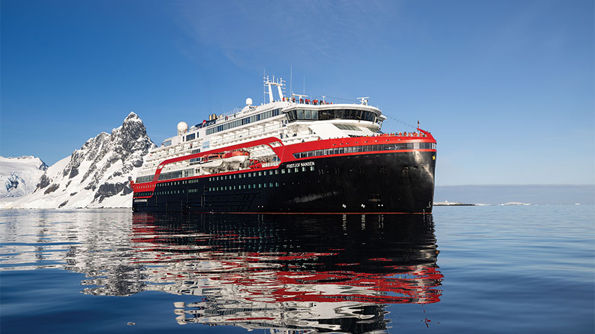 The MS Fridtjof Nansen expedition ship floats on calm blue water near snow-covered mountains, reflecting its red and black hull.
