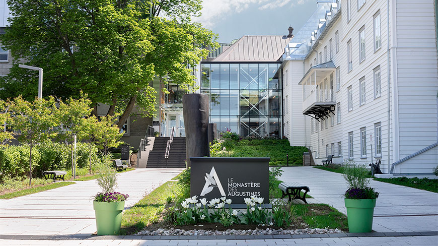 The entrance courtyard of Le Monastère des Augustines in Quebec, featuring a branded sign, historic white buildings, and modern glass architecture.