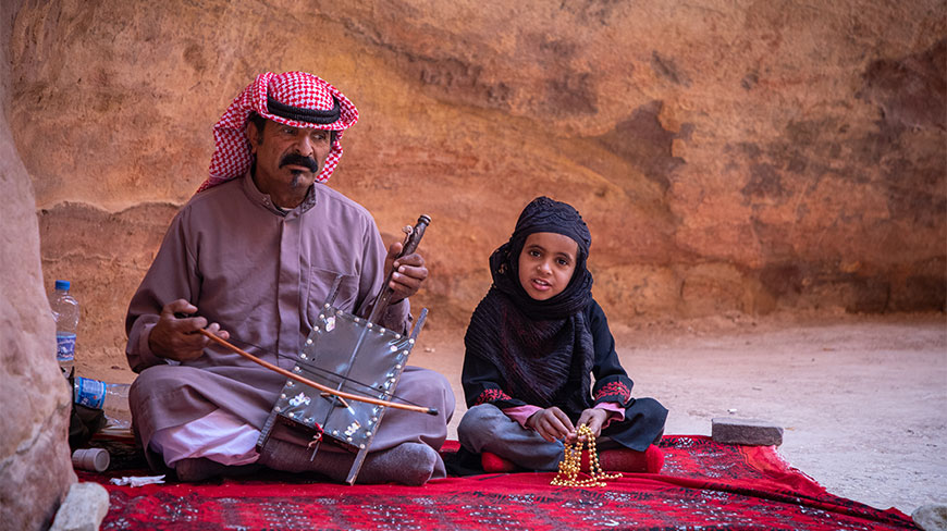 A man plays a traditional stringed instrument beside a child while resting on a rug during a Petra walking tour.