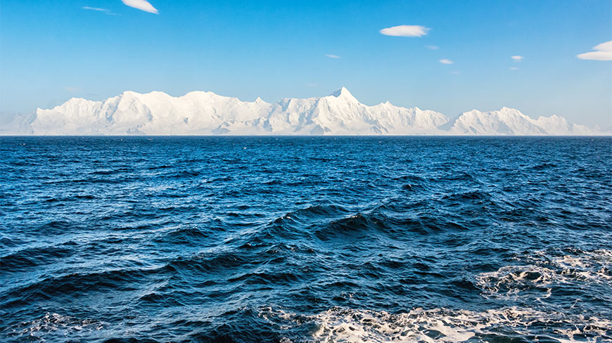 Snow-covered mountains of the South Shetland Islands rise above the choppy blue waters of the Drake Passage under a clear sky.