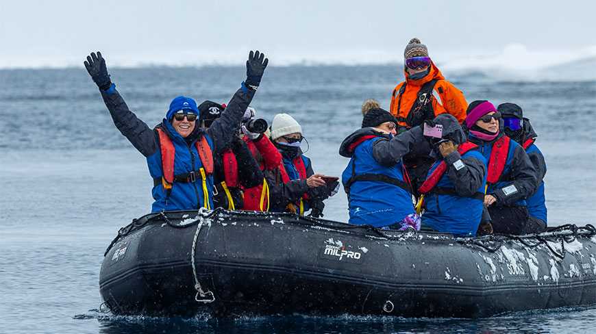 Tourists in winter gear go Zodiac cruising near Enterprise Island in Antarctica, with one person cheering and raising their arms in the air.