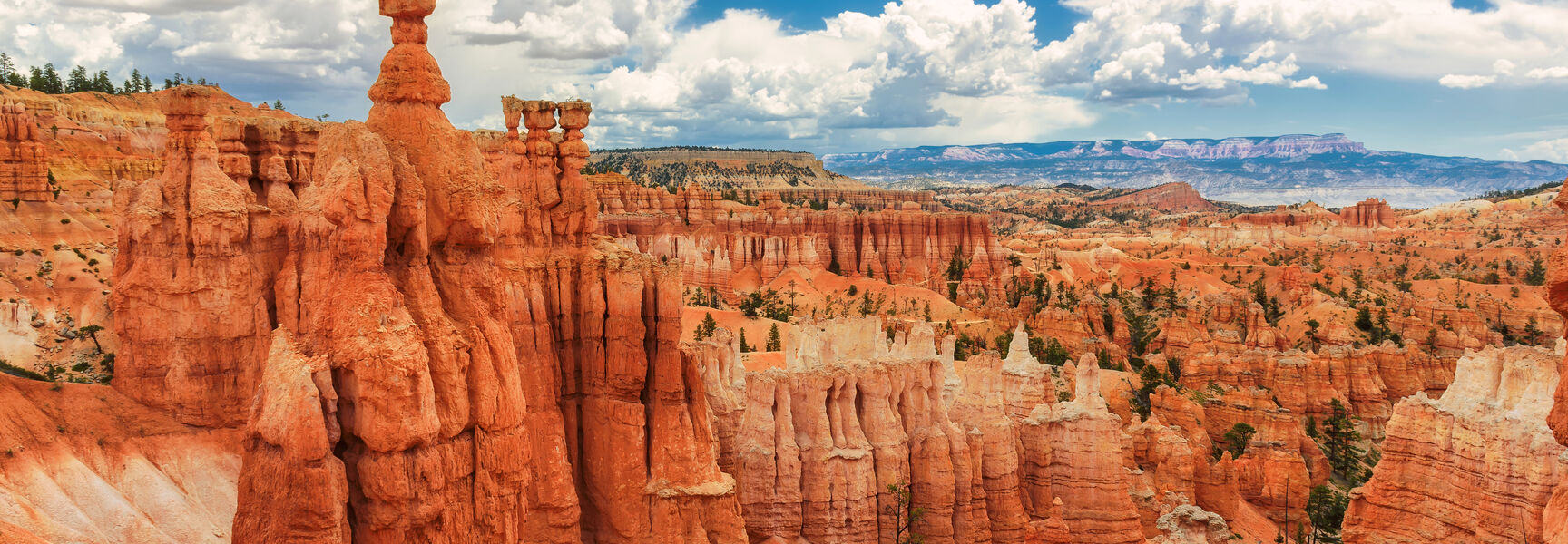 Vibrant orange hoodoos and rock spires fill the landscape of Bryce Canyon National Park in Utah under a sunny, cloud-filled sky.