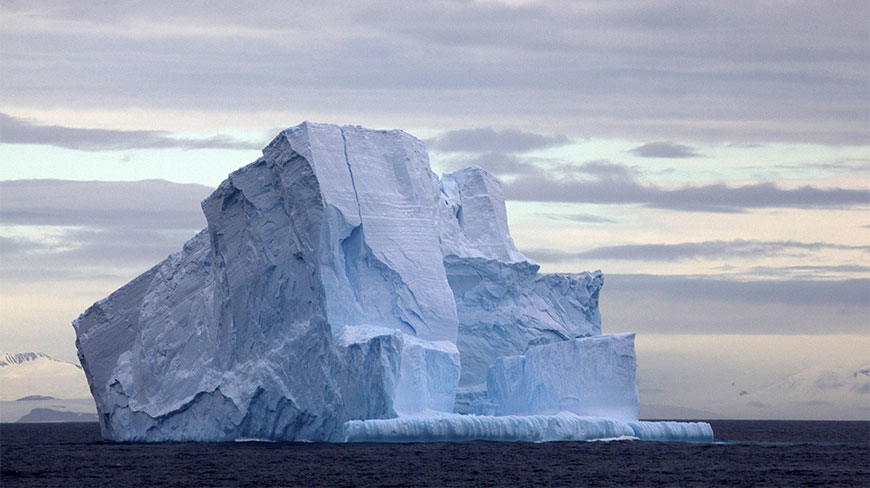 A large, jagged blue iceberg floating in the Drake Passage under a cloudy sky with distant snowy mountains.