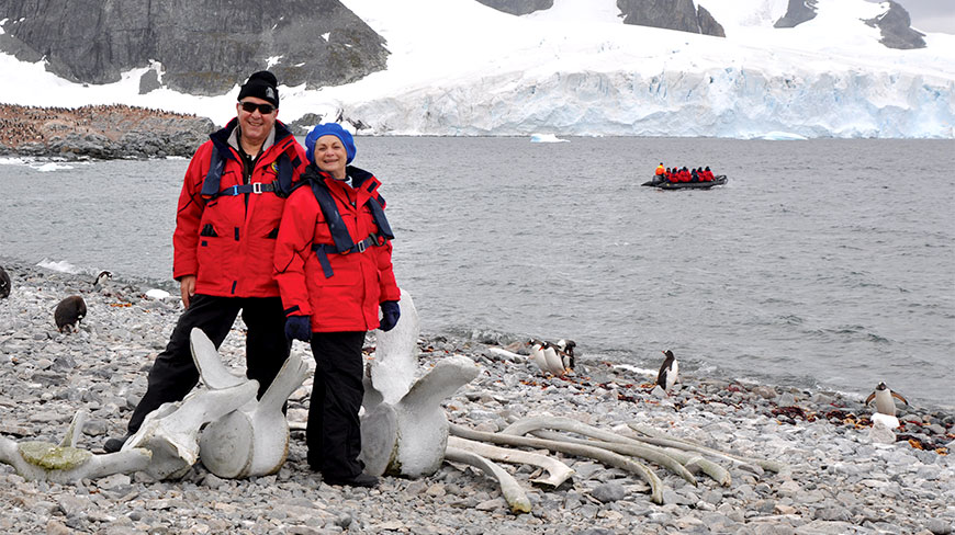 A couple wearing red parkas stands on a rocky Antarctic beach next to whale bones, with penguins and a glacier in the background.