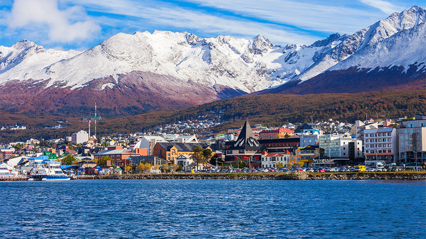 The coastal town of Ushuaia in Argentina sits below massive snow-capped mountain peaks under a clear blue sky.