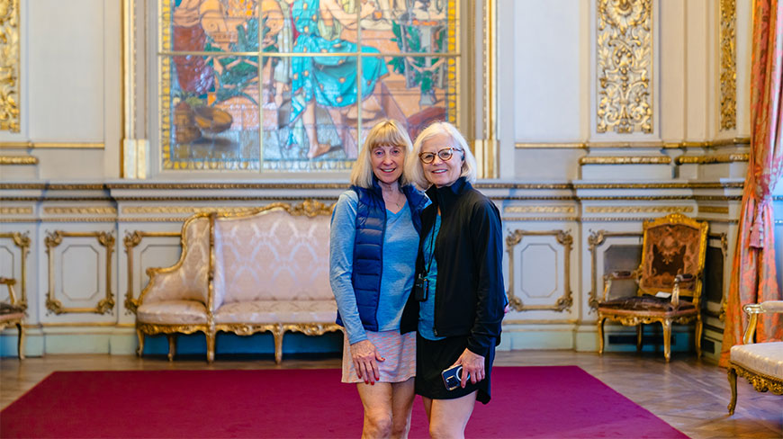Two women pose together in the ornate, gilded interior of the Teatro Colon in Buenos Aires, featuring a large stained-glass window in the background.