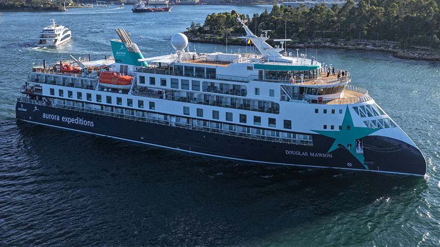 Full exterior view of the Aurora Expeditions ship Douglas Mawson afloat in the water near a lush green coastline.