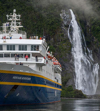 The Heritage Adventurer cruise ship sails past a massive waterfall cascading down a lush green cliff in New Zealand.