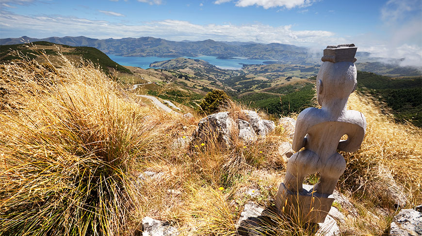 A wooden Maori God statue overlooks the scenic Akaroa Harbour and rolling green hills under a bright blue sky in New Zealand.