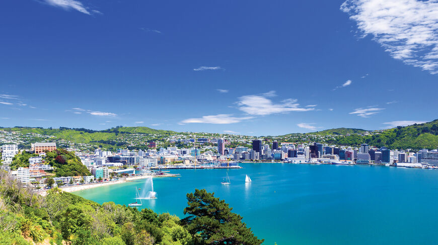 A panoramic view of Wellington Bay, New Zealand, featuring the city skyline, sailboats in turquoise water, and green hills under a bright blue sky.