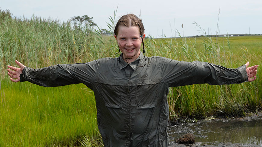 A smiling participant covered in mud stands with arms outstretched while exploring a salt marsh on Wallops Island, Virginia, during an Ocean Science Adventure.