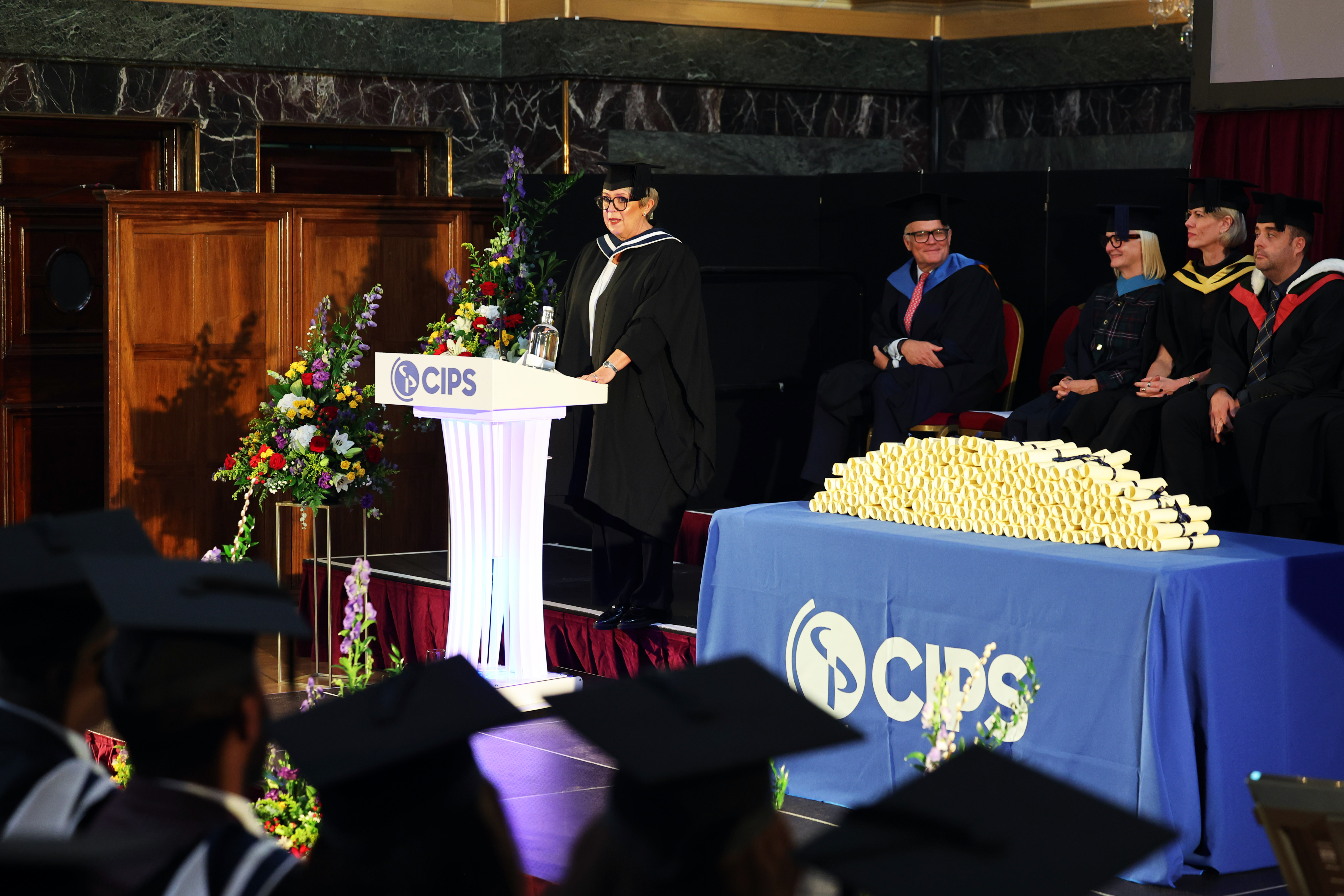 Speaker on stage giving a speech, table shows a pile of rolled up diplomas on display