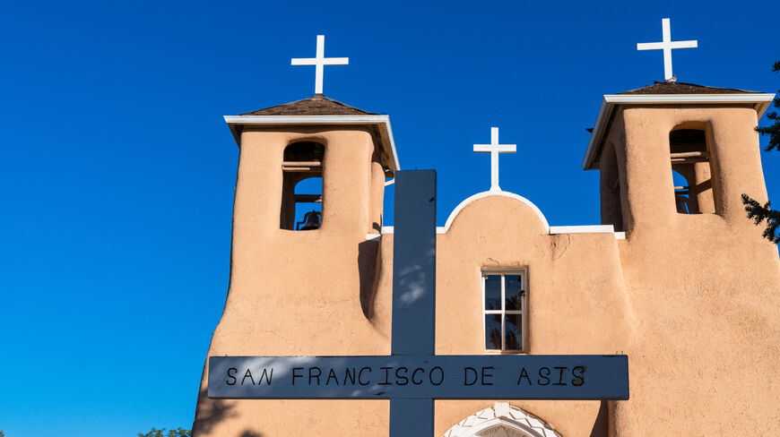 The adobe San Francisco de Asis Mission Church near Santa Fe with two bell towers and a white cross in front under blue sky.
