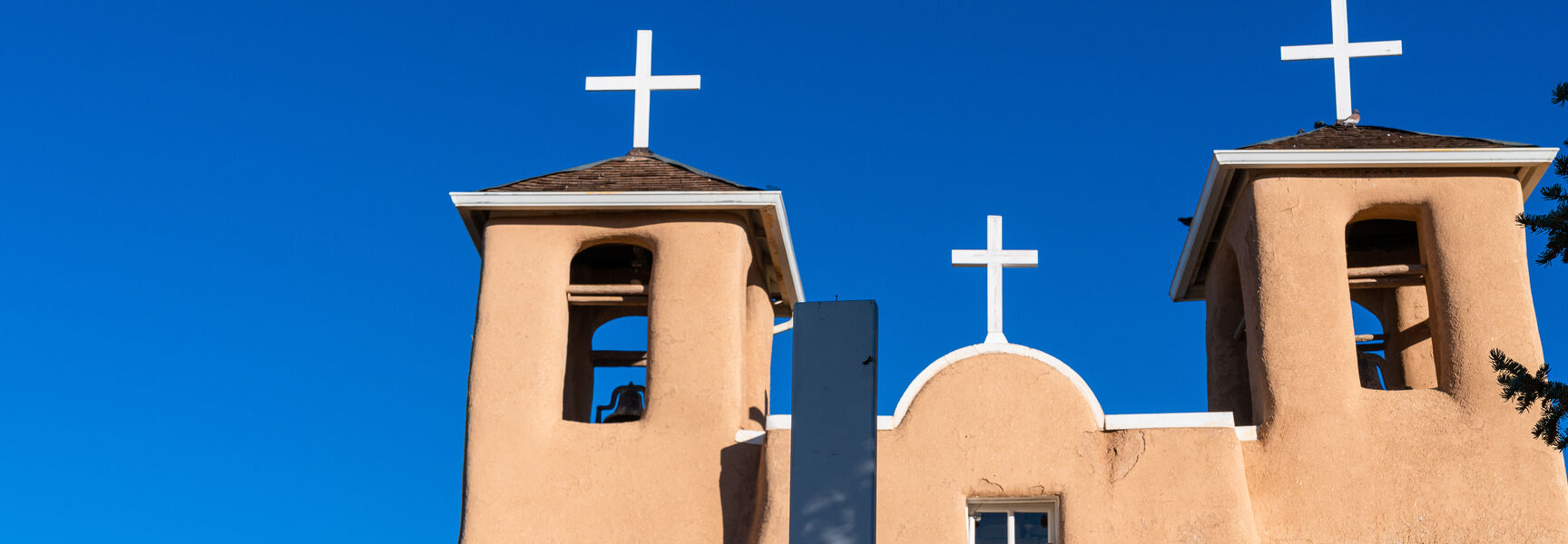 The adobe San Francisco de Asis Mission Church near Santa Fe with two bell towers and a white cross in front under blue sky.