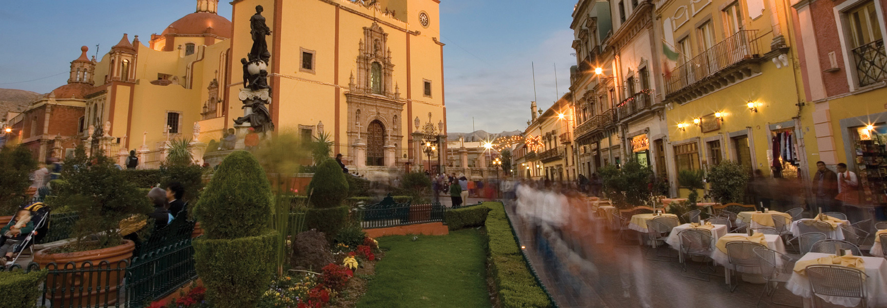 A bustling historic plaza in Mexico at dusk, with a grand church on one side and outdoor cafes on the other.