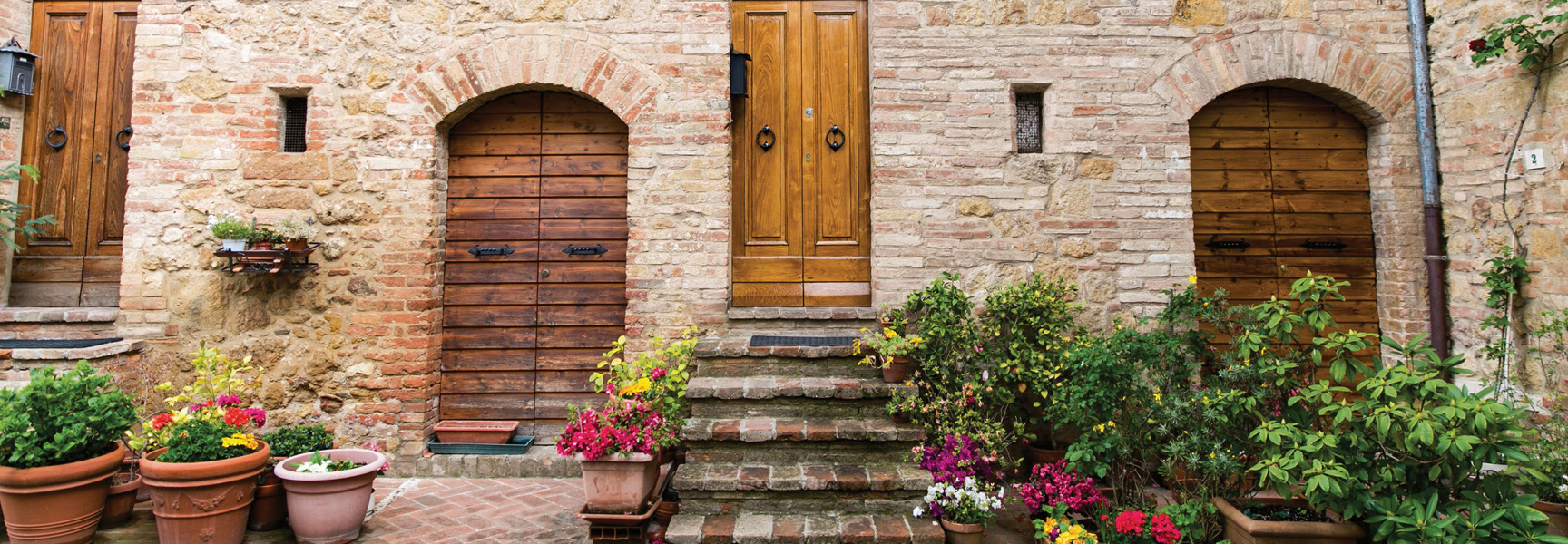 A rustic stone building in Italy with arched wooden doors, with its stone steps and entryway surrounded by colorful potted flowers.