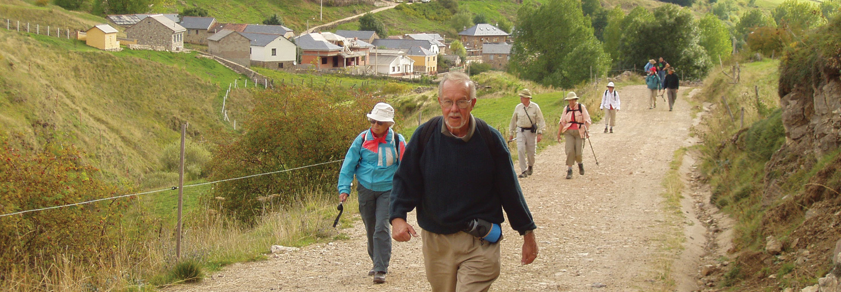 A group of senior hikers walk a dirt trail on the Camino de Santiago, with a village visible in the green rolling hills of Spain.